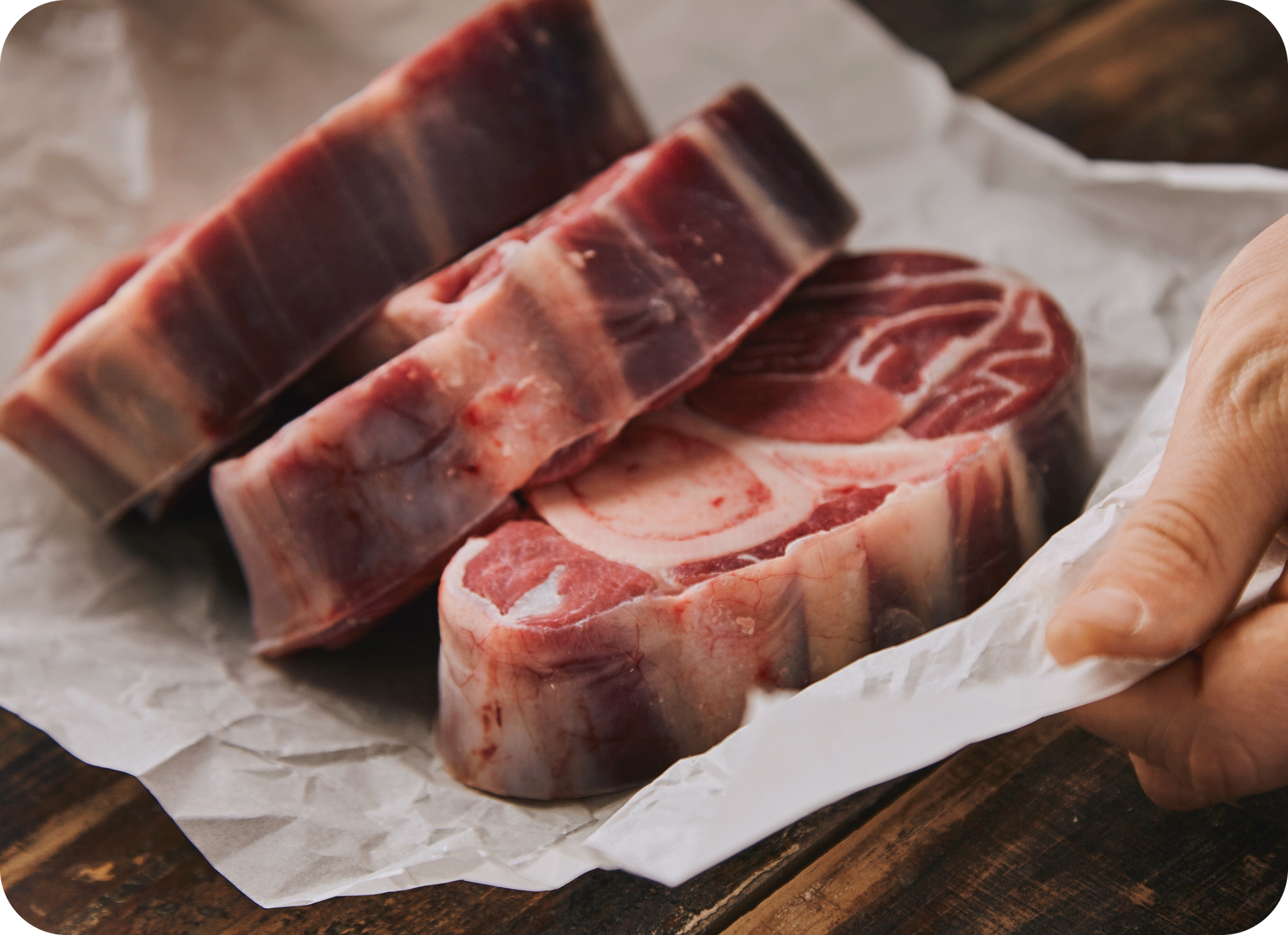 closeup-hand-holds-white-craft-paper-with-three-raw-great-leg-steaks-with-bone-it-everything-old-wooden-brushed-table 1